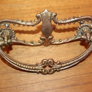 May include: Ornate brass drawer pull with a decorative, scrolled design. The pull has a curved shape and two decorative knobs on either side.