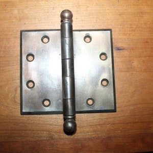 May include: A silver door hinge with a rounded top and bottom. The hinge has eight holes for screws and is attached to a wooden surface.