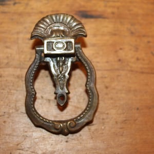 May include: A vintage brass drawer pull with an ornate design. The pull features a decorative top with a fan-shaped design and a ring-shaped handle.