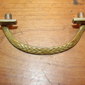 May include: A brass drawer pull with a decorative woven design. The pull has a square base and a curved handle.