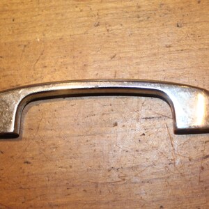 May include: A vintage-style, silver-toned cabinet pull handle. The handle has a curved design with rounded ends and a rectangular base. The metal has a slightly aged appearance, suggesting it is an antique or a reproduction.