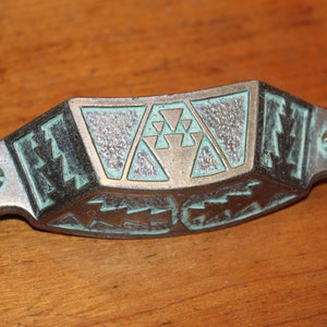 May include: A vintage, green and silver metal cabinet pull with a geometric design. The pull has a curved shape and two screw holes.