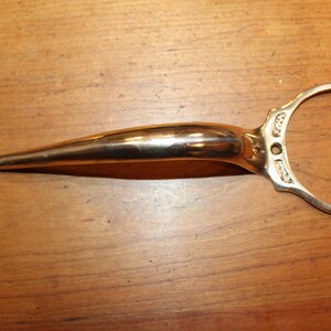 May include: A gold-colored door handle with a diamond-shaped end and a circular handle. The handle has decorative detailing and is likely made of metal. The handle is shown on a wooden surface.