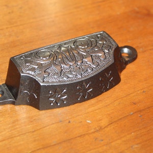 May include: A silver-colored metal drawer pull with an ornate floral design. The pull has two holes for mounting.