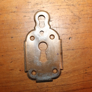 May include: A brass keyhole cover with a decorative design. The cover has a keyhole in the center and four small holes around the edges.