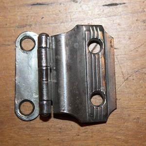 May include: A silver and brass hinge with a decorative design on the brass side. The hinge has three holes on each side for screws.