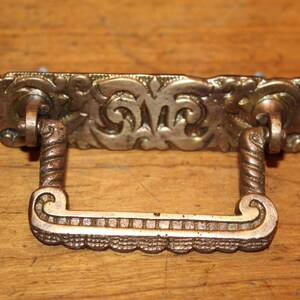 May include: Ornate brass drawer pull with a decorative, scrolled design. The pull has a rectangular shape with a raised, textured center.