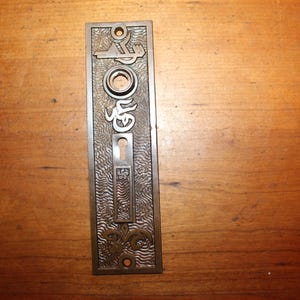 May include: A vintage brass door plate with an ornate design and a keyhole. The plate has a textured surface and is rectangular in shape.