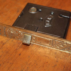 May include: A brass door latch with a decorative design and a black metal lock mechanism. The lock mechanism has the word "ELEY" printed on it.