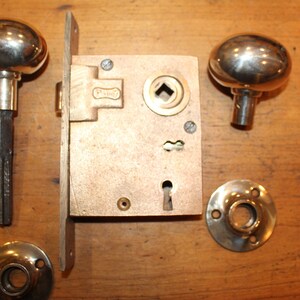 May include: A vintage door lock set with two round, chrome door knobs, a brass lock plate, and two chrome escutcheons. The lock plate has a keyhole and a square-shaped hole for a door handle.