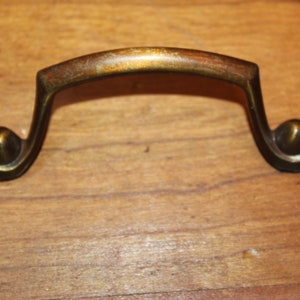 May include: A vintage brass drawer pull with a curved handle and decorative ball-shaped ends. The hardware has a weathered appearance, suggesting age and use. The pull is designed for furniture restoration or as a decorative accent.