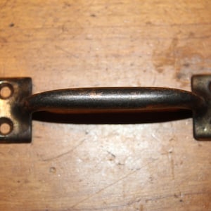 May include: A vintage-style metal handle with a curved grip and rectangular mounting plates. The handle is dark bronze with a slightly worn appearance, and each plate has two screw holes. This hardware is suitable for cabinets or drawers.