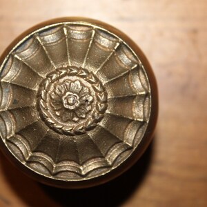 May include: A brass door knob with a decorative, ornate design. The knob features a central flower-like motif surrounded by a series of concentric circles and radiating lines.