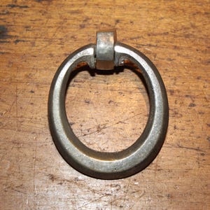 May include: Oval-shaped, antique metal drawer pull with a loop handle and a rectangular mounting bracket. The hardware has a tarnished, aged appearance, suggesting vintage or rustic decor. The pull is set against a wooden surface.