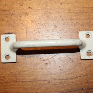 May include: A vintage, off-white metal handle with two rectangular mounting plates, each with two holes. The handle is a curved bar. The item is set against a wooden surface.