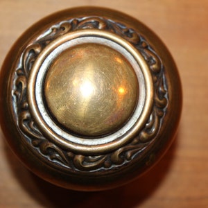 May include: A round, brass door knob with an ornate, detailed design. The knob has a raised, circular center.
