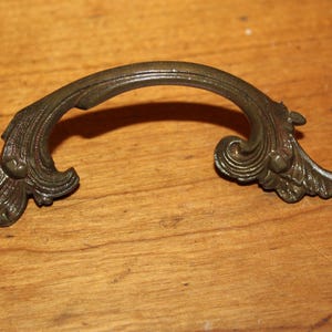May include: A decorative, ornate, bronze-colored metal drawer pull with a curved shape and intricate floral designs.