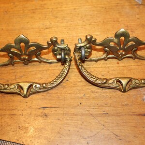 May include: Two antique brass drawer pulls with ornate floral and scrollwork designs. The pulls have a curved handle and a decorative top piece. The hardware is a warm gold color and is set against a wooden surface.