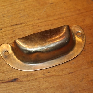 May include: A brass-toned, half-moon shaped drawer pull with two mounting holes. The hardware has a curved, cup-like design and a slightly aged appearance. The pull is set against a wooden surface.