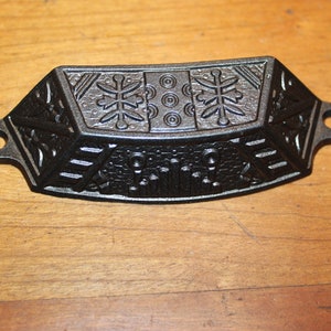 May include: Black cast iron drawer pull with a decorative design featuring geometric patterns and stylized snowflakes. The pull has two holes for mounting.