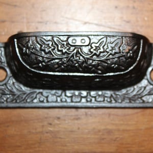 May include: Black cast iron drawer pull with a floral design. The pull has a rectangular base with rounded edges and a curved top. The floral design features leaves and vines.