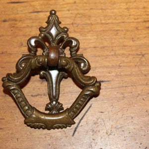 May include: Ornate brass drawer pull with a decorative design. The pull has a ring shape with a detailed center piece.