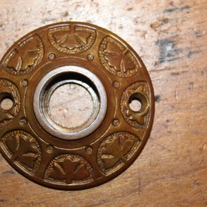 May include: A round, brass door knob escutcheon with a decorative design of leaves and flowers. The escutcheon has a hole in the center for the knob.