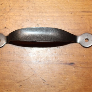 May include: A dark, metallic handle with a curved center and circular ends, each with a hole. The handle is likely for a drawer or cabinet, and is set against a wooden surface.