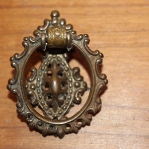 May include: Antique brass drawer pull with an ornate, oval design. The pull features a decorative ring and a detailed central element. The hardware is set against a wooden surface, highlighting its vintage aesthetic. Ideal for furniture restoration or adding a classic touch.