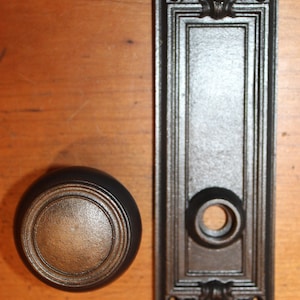 May include: A black cast iron door knob and backplate with ornate detailing. The knob has a round, slightly domed shape with a smooth, slightly textured surface. The backplate has a rectangular shape with a decorative design at the top and bottom.