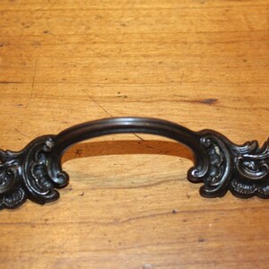 May include: Ornate, dark bronze cabinet pull with a curved handle and decorative floral scrollwork on each end. This hardware is designed for furniture and adds a vintage touch.