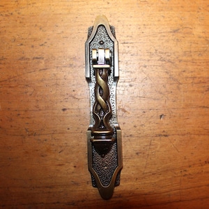 May include: A decorative metal door knocker with a twisted design and a pointed top. The knocker is finished in a brass color.