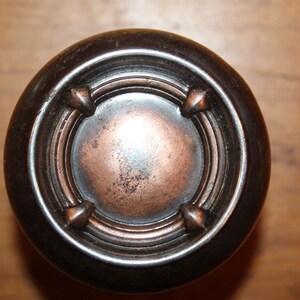 May include: A close-up of a round, dark brown door knob with a copper-colored center. The knob has a decorative design with four raised points around the center.