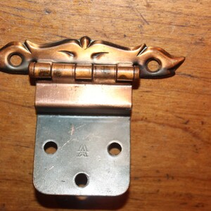 May include: A vintage copper and silver door hinge with a decorative, ornate top. The hinge has a small 'A' stamped on the bottom.