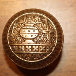 May include: A detailed, antique-style, gold-colored knob with an ornate design. The circular knob features a central motif of a vase and floral elements, surrounded by geometric patterns. The knob is likely made of metal and is set against a wooden surface.