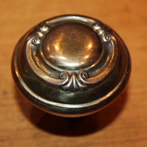 May include: A round, brass door knob with an ornate design. The knob has a raised, circular center and a decorative border.
