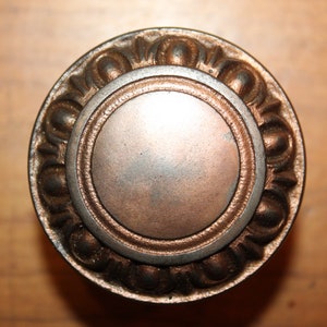 May include: A round, ornate door knob with a silver center and a bronze outer ring. The knob has a decorative pattern of raised circles around the edge.