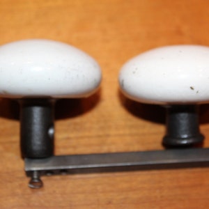 May include: Two white porcelain door knobs with black metal stems and a metal bar connecting them.