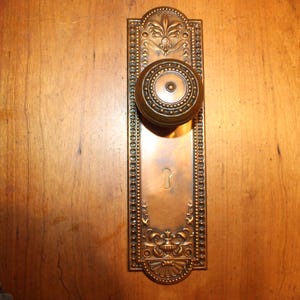 May include: Ornate antique door hardware featuring a brass doorknob and backplate. The backplate has decorative floral and beaded detailing, with a keyhole. The doorknob is round with concentric circles.