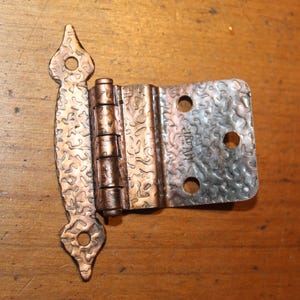 May include: A copper-toned metal cabinet hinge with a textured, hammered surface finish. The hinge features a decorative, elongated plate with two screw holes and a rectangular plate with three screw holes. The word JAPAN is stamped on the metal.