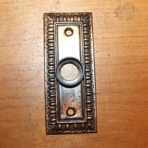 May include: A vintage-style door knocker with a decorative, ornate frame. The knocker is made of metal and has a dark brown finish.