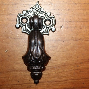 May include: A decorative, ornate, black metal cabinet pull with a silver finish. The pull has a bell shape with intricate details and a ring at the top.