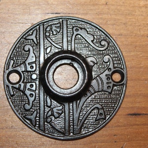 May include: A round, black metal door knob with an ornate design. The knob has a floral pattern and a small hole in the center.