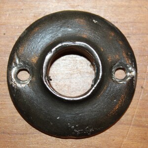 May include: A black metal door knob rosette with two holes for screws. The rosette has a slightly worn, aged look.