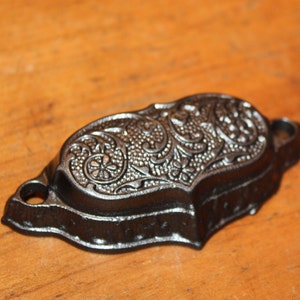 May include: A black cast iron drawer pull with an ornate floral design. The pull has a curved shape and two holes for mounting.