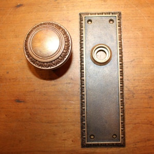 May include: A set of antique brass door hardware, including a round door knob with a decorative border and a rectangular backplate with a keyhole and two holes for mounting.