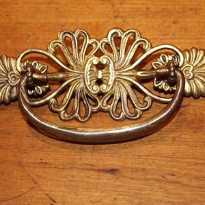 May include: Ornate brass drawer pull with a curved handle and decorative floral and scrollwork details. The hardware is a warm gold color and is mounted on a wooden surface.