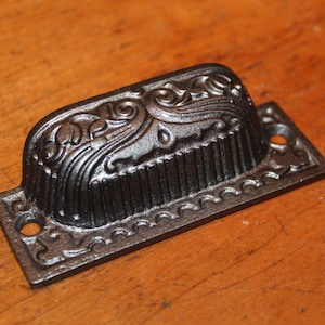 May include: Black cast iron drawer pull with an ornate design. The pull has a rectangular base with a raised, curved top.