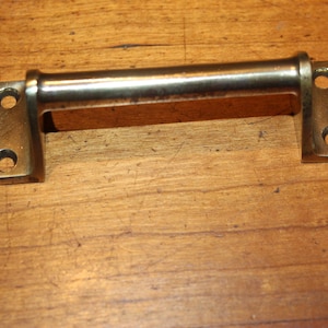May include: A brass-toned cabinet pull handle with a rectangular bar and two mounting plates, each with two screw holes. The handle is set against a wooden surface.