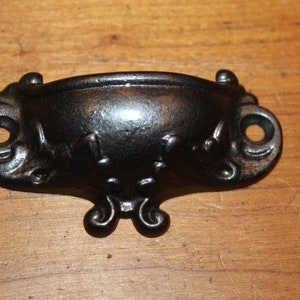 May include: Black metal drawer pull with ornate details and a curved shape.